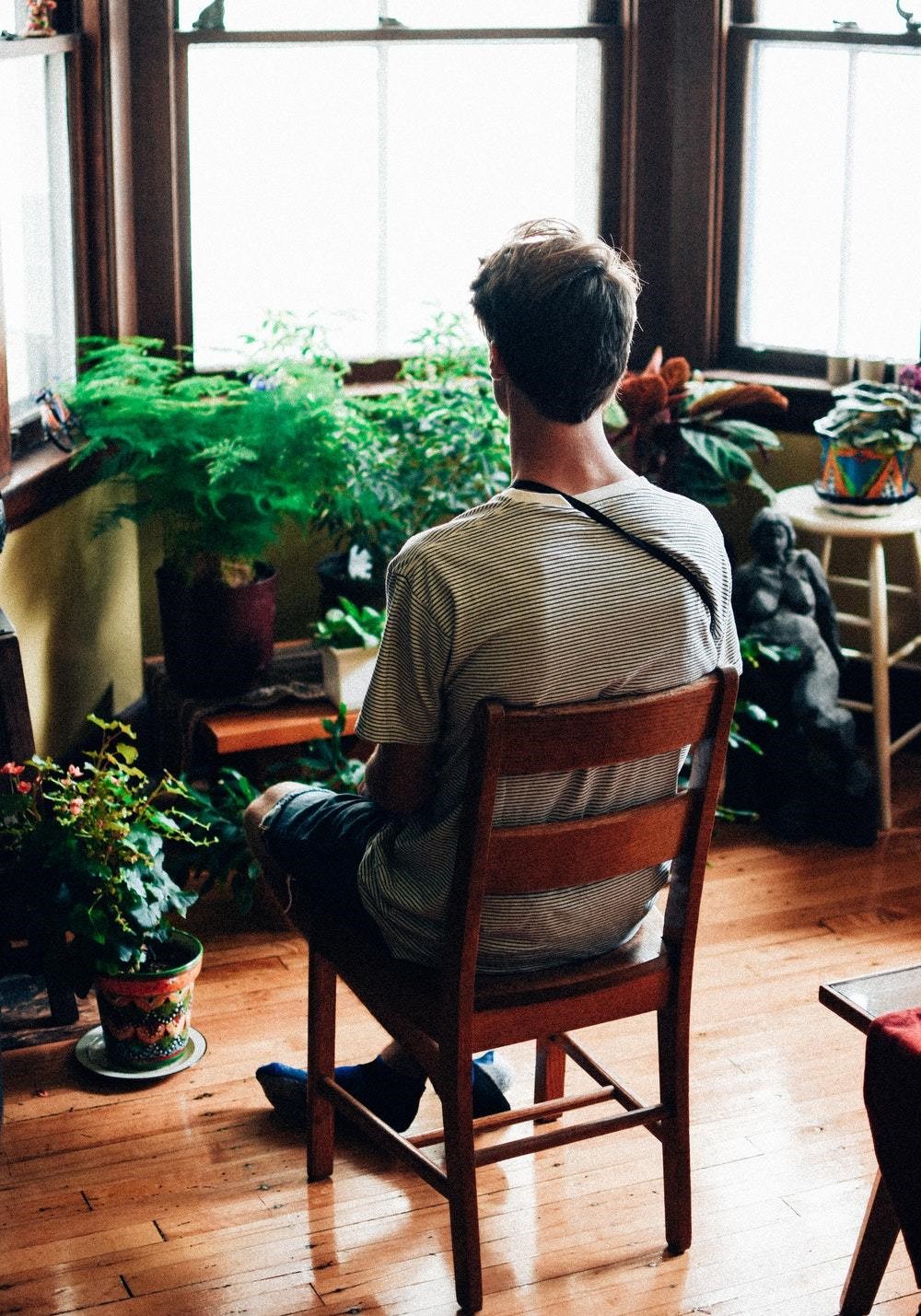 main in chair indoors looking at plants along a window seal